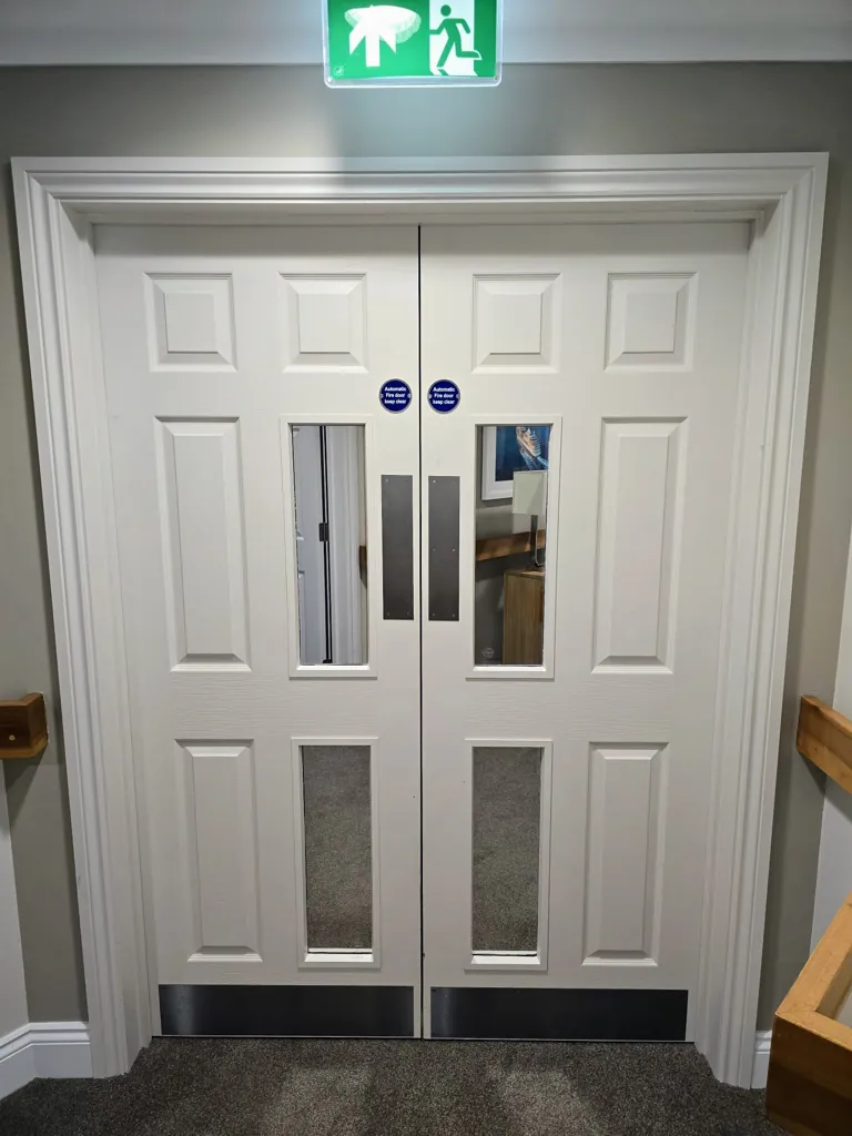 White double fire exit doors with narrow vision panels and illuminated exit sign above