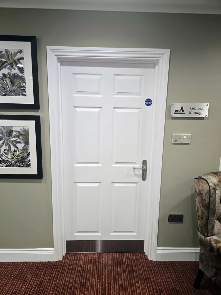 White panel door with metal kick plate and room signage in a furnished care home interior