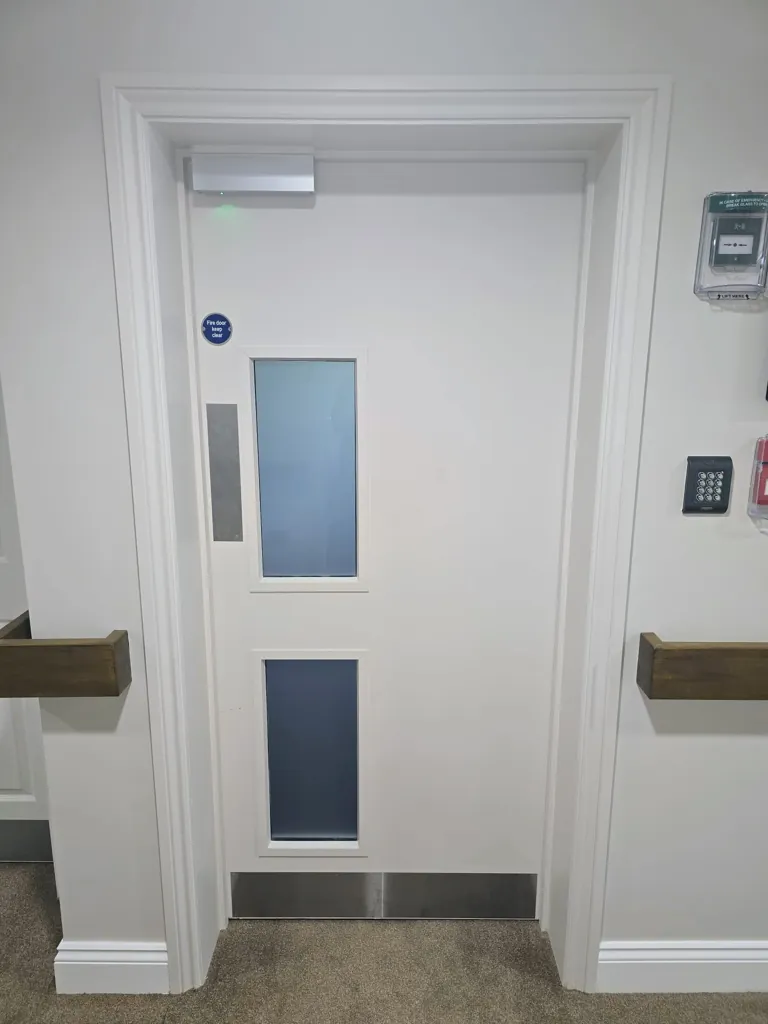 White fire door with vertical vision panels, overhead closer and access keypad on wall