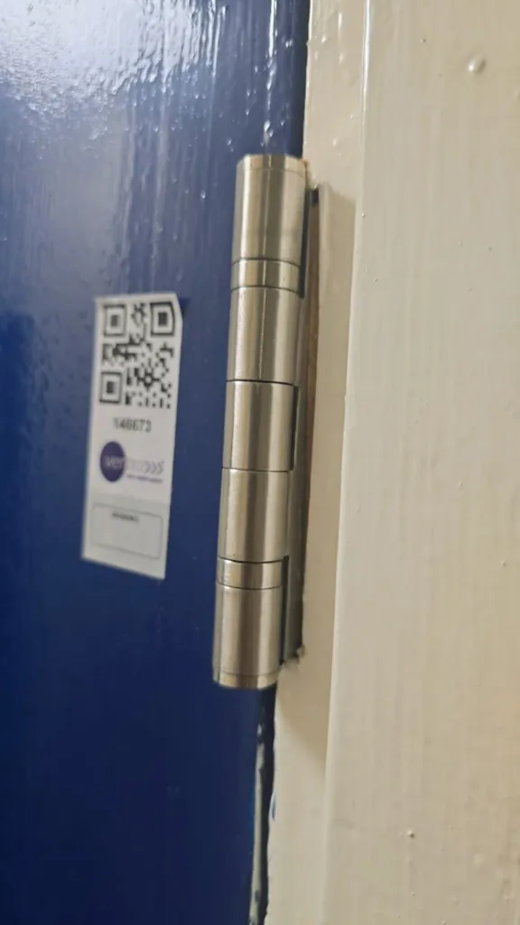 Close-up of fire door hinge on blue door frame with QR code inspection label