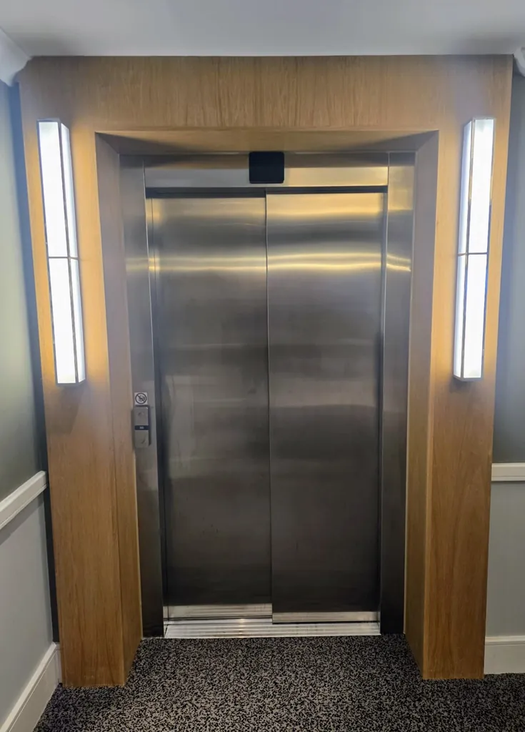 Stainless steel lift doors set within wood panelled surround with vertical wall lights